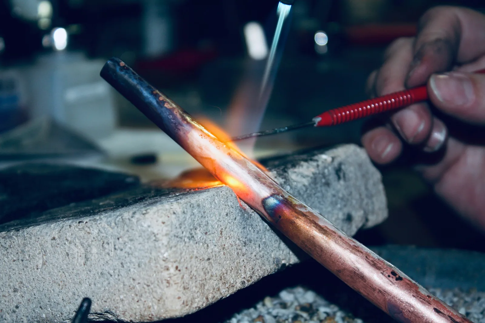 A torch flame heating a joint in a copper line on a fire brick, one hand holding a pick.